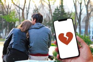 A couple sitting in a forest with the woman leaning her head on the man's shoulder and an app on a phone showing a broken heart.